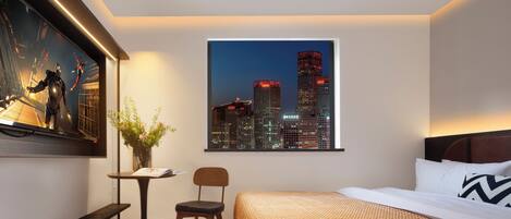 A modern hotel room with a cityscape view, a bed with a patterned bedspread, a small table with a chair, and a framed picture on the wall.