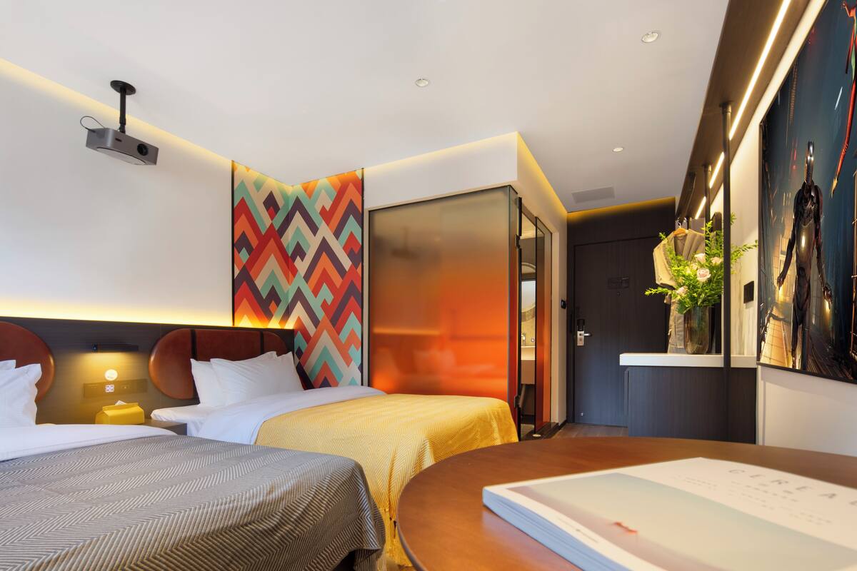 A hotel room with two beds, a desk, a projector, a colorful wall, and a large window.