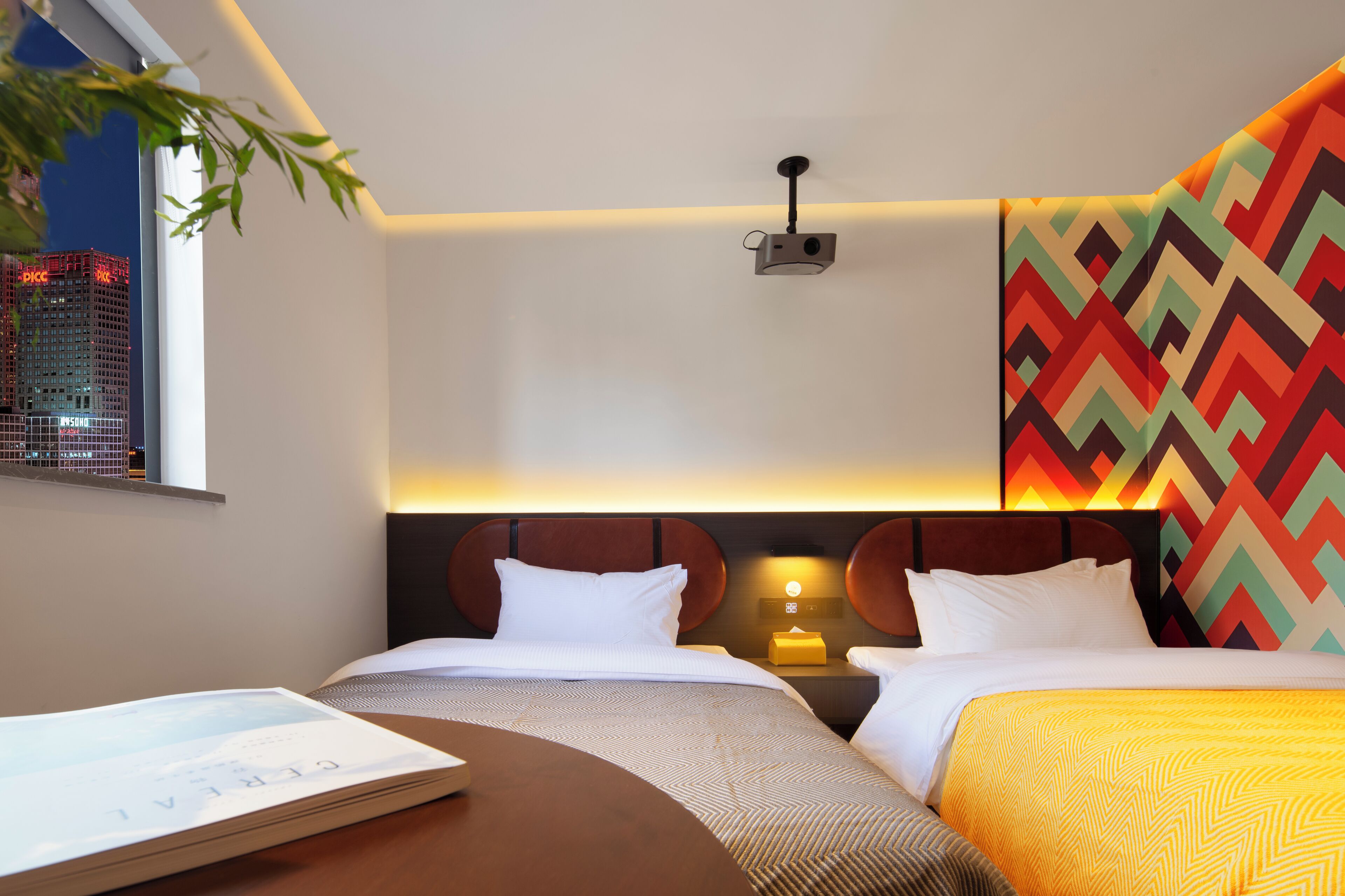 Een hotelkamer met twee bedden, een nachtkastje met lamp, een raam met uitzicht op de stad en een muur met een geometrisch patroon.