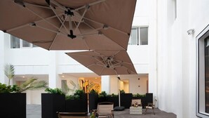 An outdoor seating area with wooden flooring, black planters with greenery, and beige umbrellas.