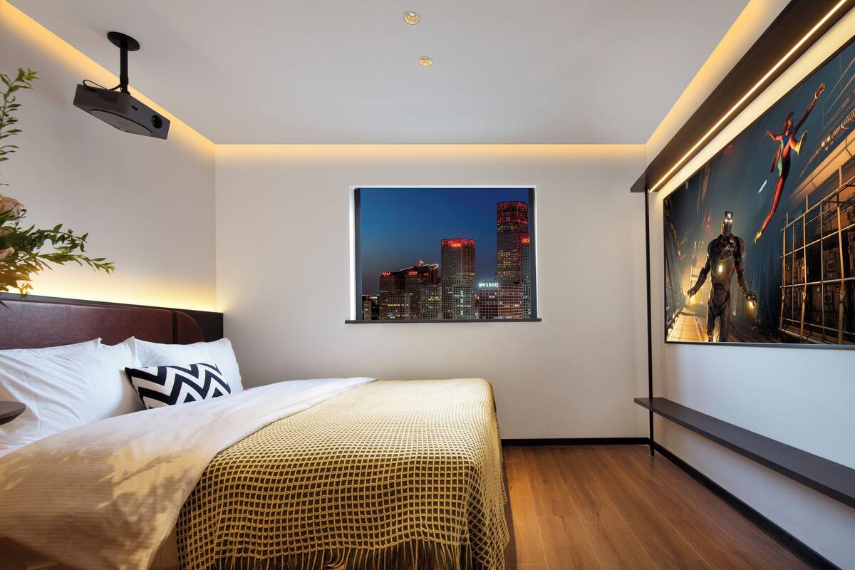 A modern hotel room with a cityscape view, a bed with a patterned bedspread, a projector mounted on the ceiling, and a large framed artwork on the wall.