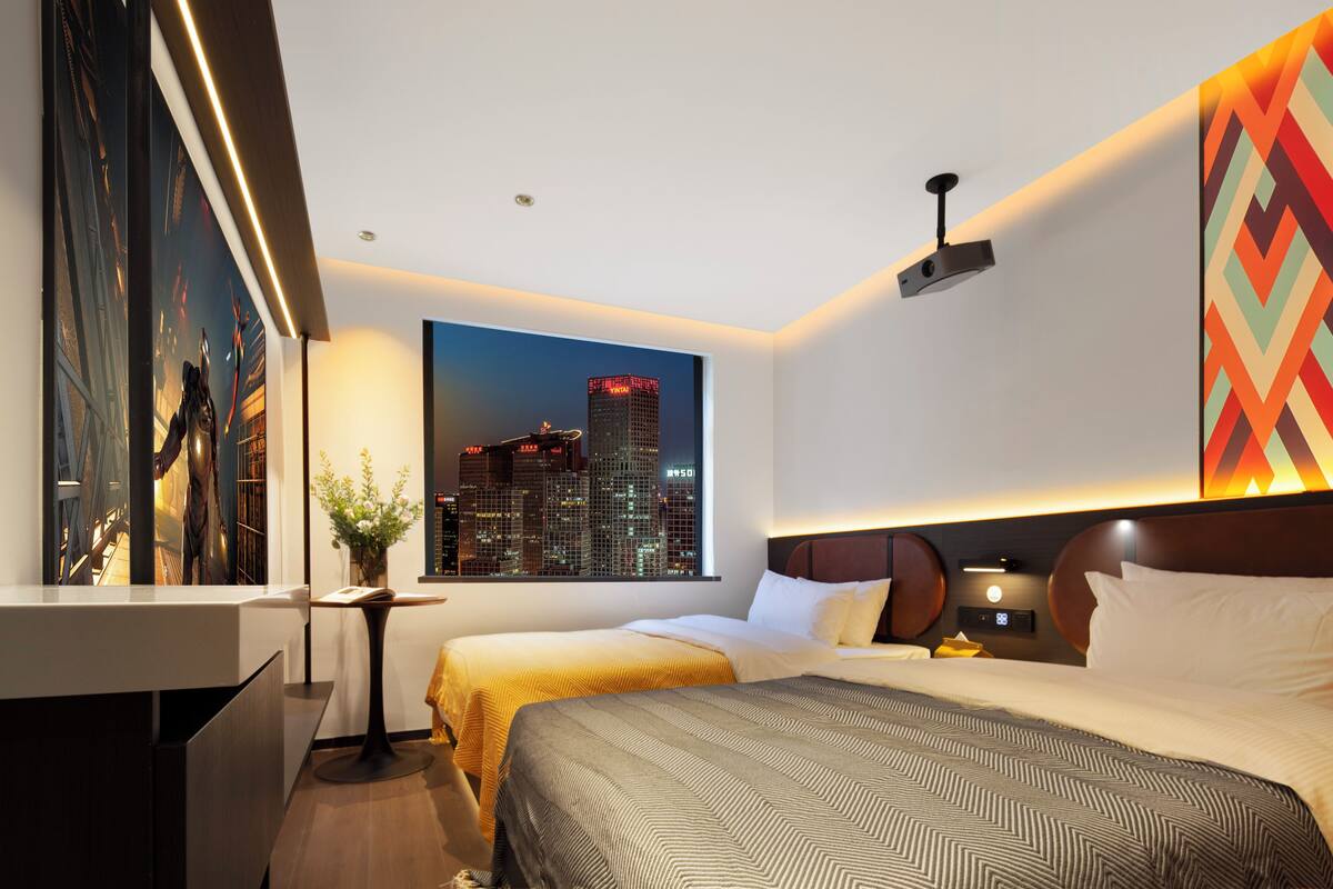 A modern hotel room with two beds, a cityscape view, a desk, and a decorative wall piece.