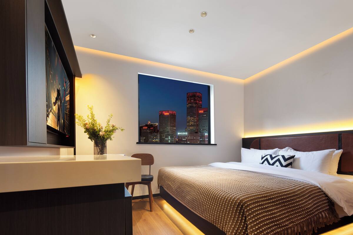 A modern hotel room with a cityscape view, a desk with a chair, and a neatly made bed.
