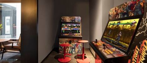 Un salón de juegos con dos máquinas recreativas, taburetes rojos y una pantalla grande mostrando un juego de lucha.