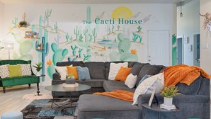 A living room with a large cactus mural, a green sofa, a gray sectional sofa, and a coffee table. - The Cacti House - Hot Tub, Game Room, Cactus Mural, Minutes from Downtown Dallas (Dallas)