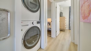 Ein Waschbereich mit zwei Waschmaschinen mit Frontlader, einem Holzkasten und einem Flur, der zu einem Schlafzimmer führt.
