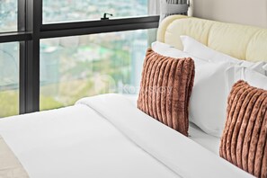 A bed with white bedding and brown pillows, a window with a view of greenery, and a yellow headboard. - Kiyo by Kozystay - Kemayoran (Jakarta)