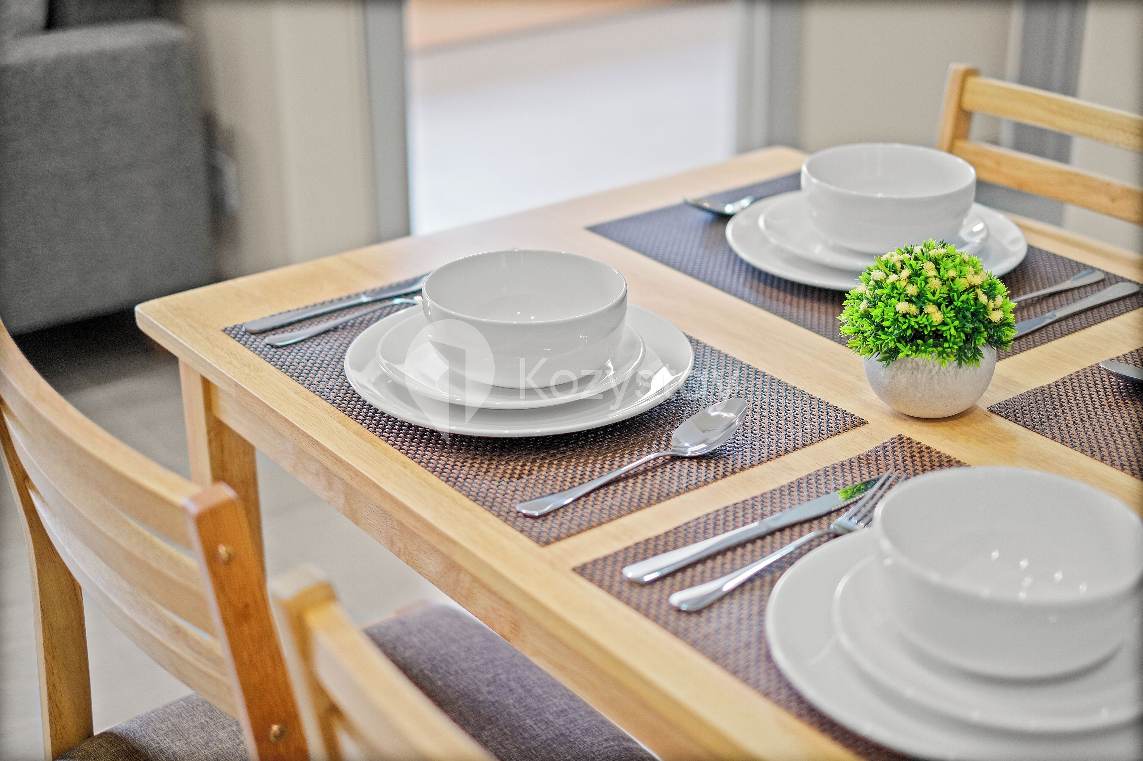A dining table set with white plates, bowls, and cutlery, a small potted plant, and a woven placemat.