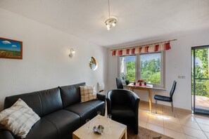 A living room with a black leather sofa, a dining table with chairs, a window with curtains, and a wall-mounted light fixture.