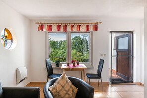 Une pièce comprenant une table à manger, deux chaises, un canapé, une fenêtre donnant sur un espace verdoyant et une porte.