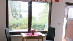 A room with a dining table, two chairs, a sofa, a window with a view of greenery, and a curtain. - Holiday Home 'Seeblick' with Lake View, Private Terrace and Private Garden (Leezen)