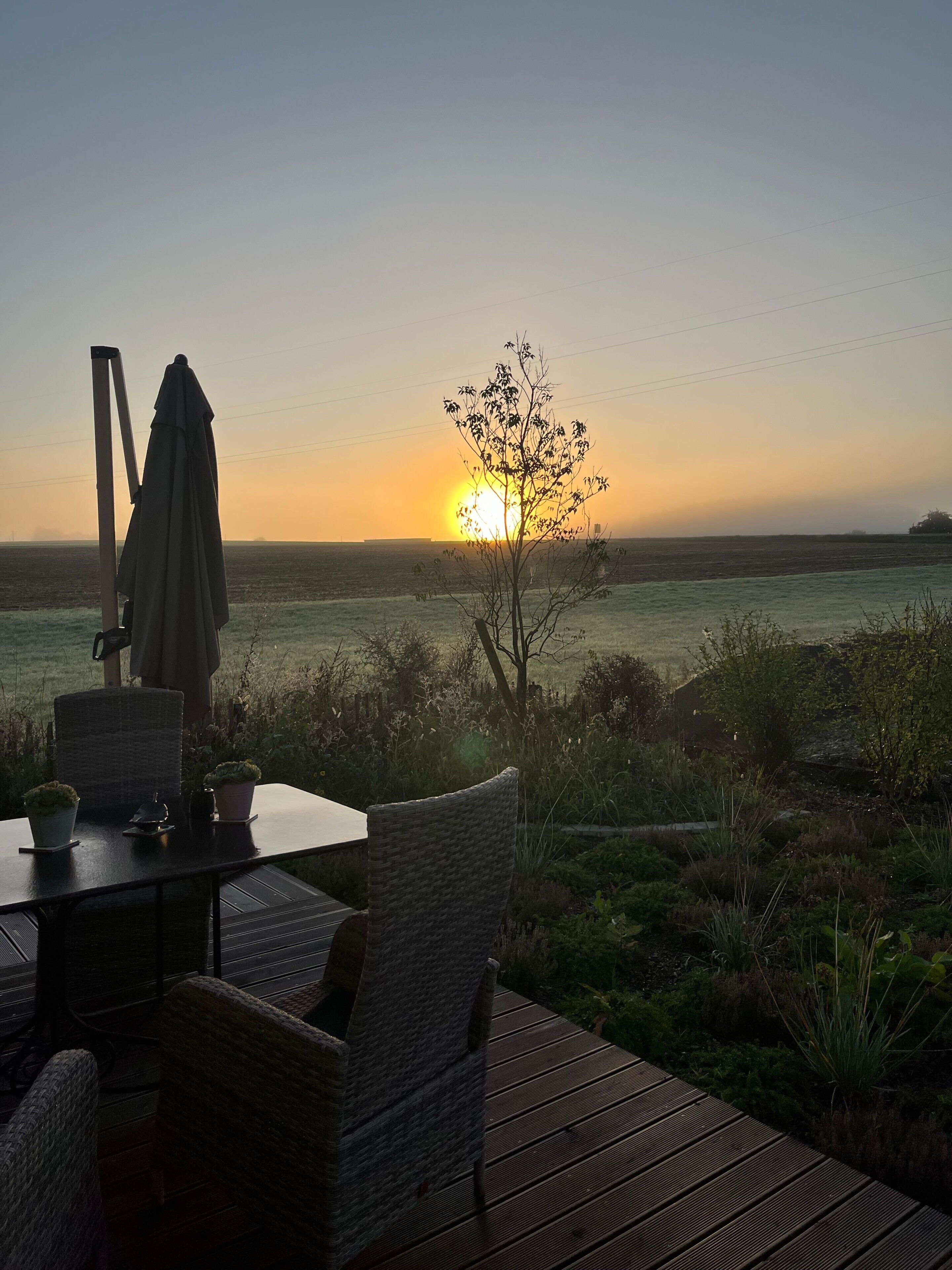 A deck with wicker furniture, a table, and a chair, overlooking a field with a sunset in the distance.