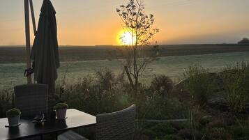 A deck with wicker furniture, a table, and a chair, overlooking a field with a sunset in the distance.