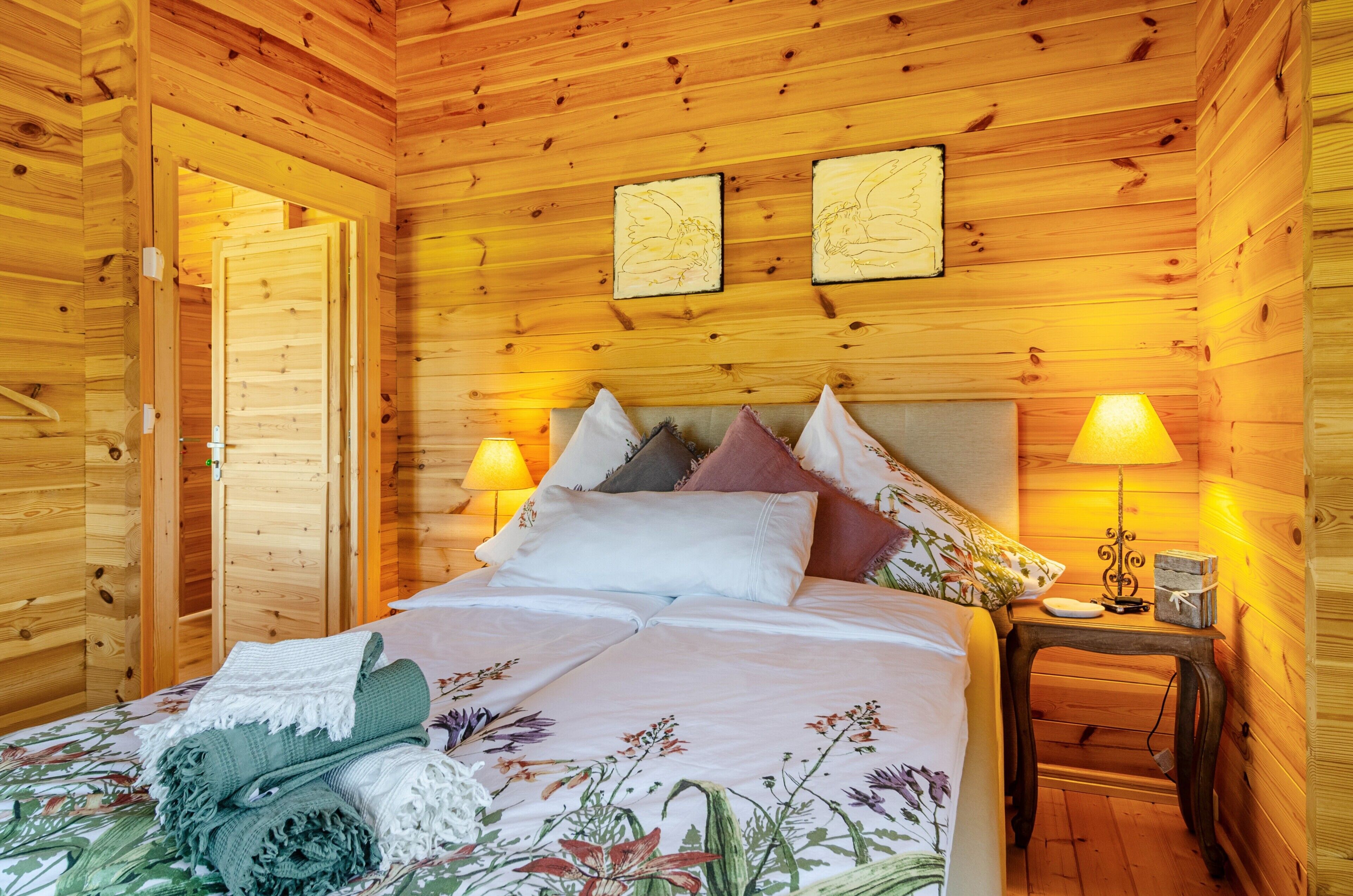 Un dormitorio de cabaña de madera con cama, almohadas, una mesita de noche y dos lámparas fijadas a la pared.