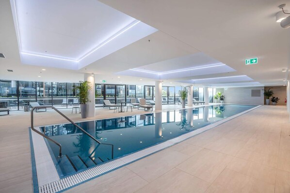 Piscina interior moderna con paredes de vidrio transparente, sillones de descanso y plantas en macetas.