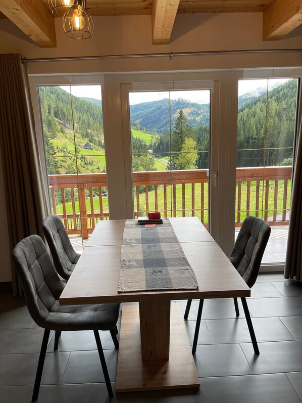 A dining area with a large table, four chairs, and a view of a mountainous landscape through large windows.