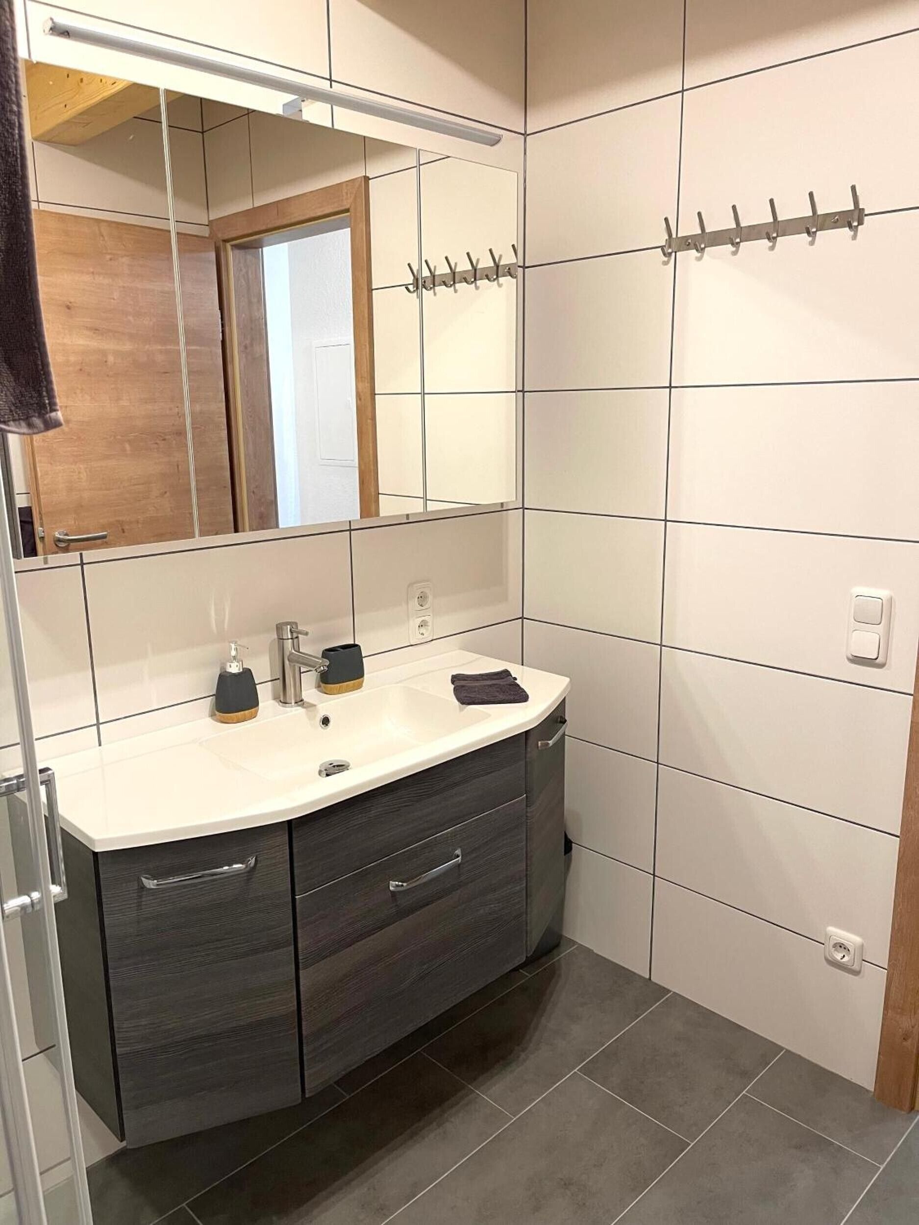 Een badkamer met een witte wasbak, een donker houten opbergmeubel, een spiegel en een handdoekhouder.