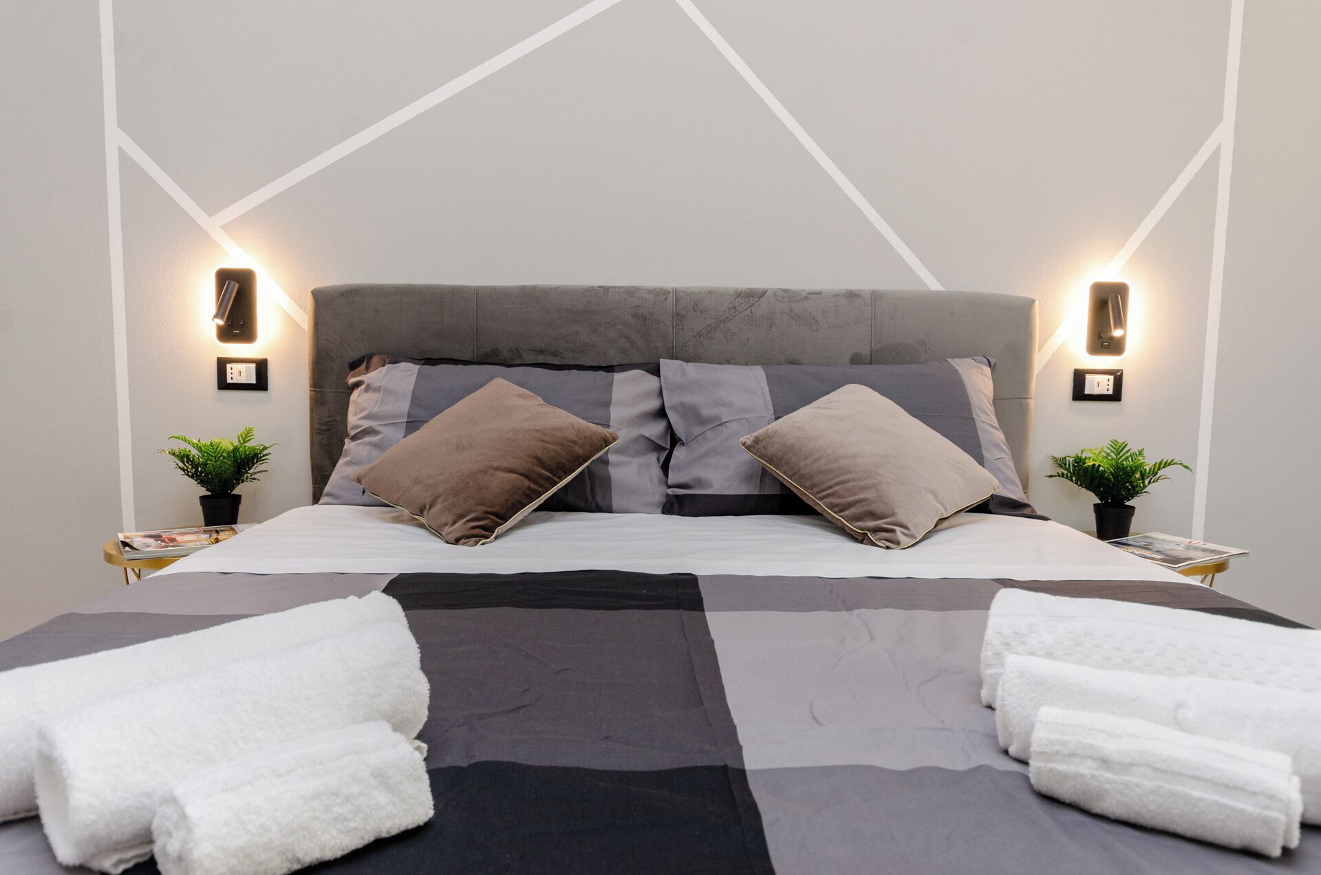 A neatly made bed with a grey and black checkered bedspread, two pillows, and two white towels on the floor.