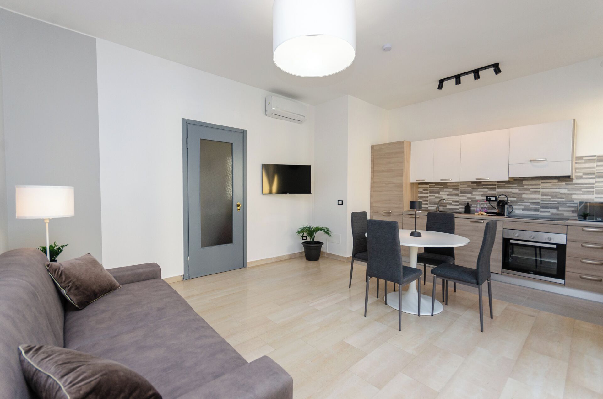 A modern living room with a grey sofa, a dining table with chairs, a TV mounted on the wall, and a kitchen area with cabinets and a stove.