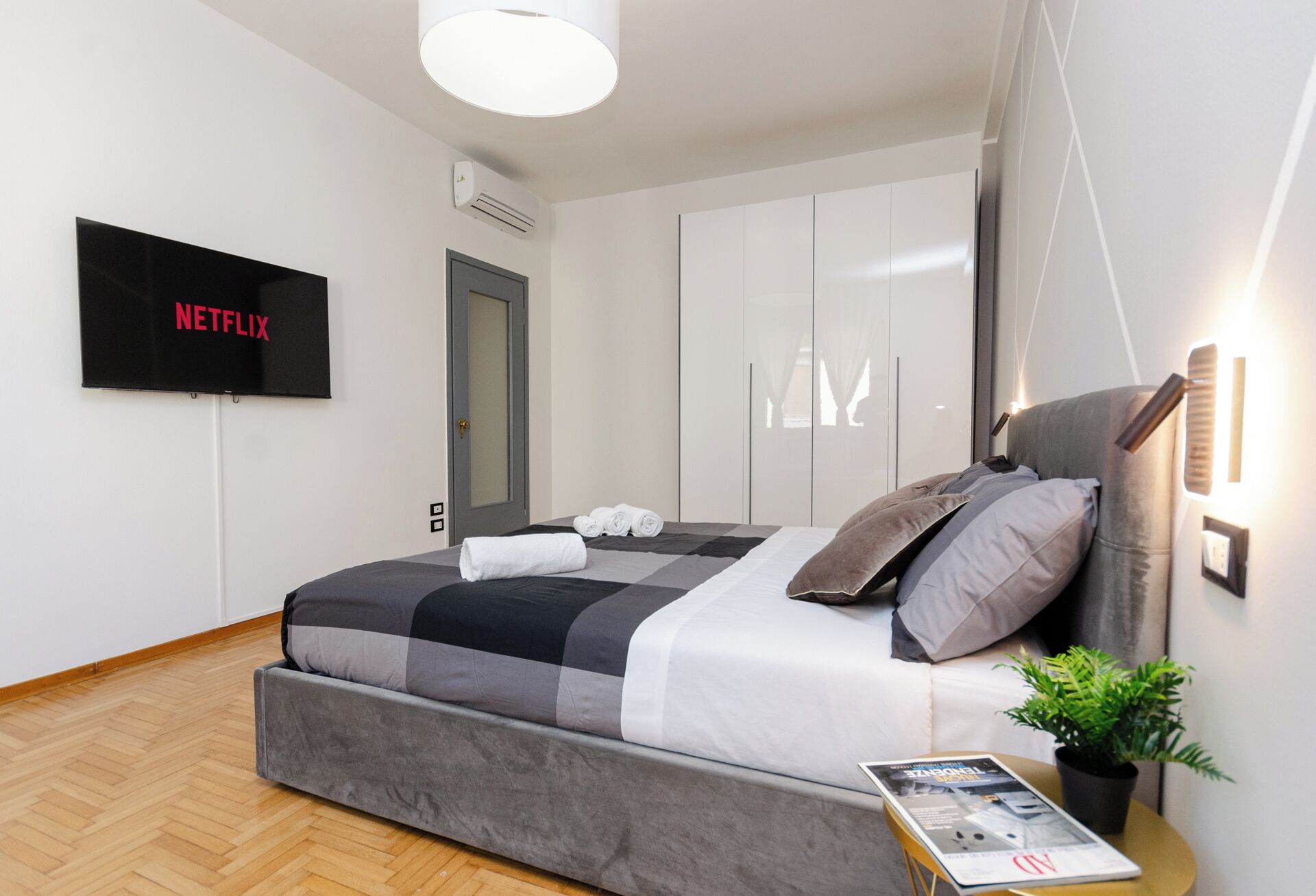 A modern bedroom with a grey bed, a flat-screen TV, and a potted plant.
