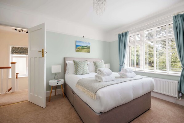 A bedroom with a large bed, a bedside table, a nightstand, a window with curtains, and a door leading to another room. - Primrose - Pet Friendly Family Home (Poole)