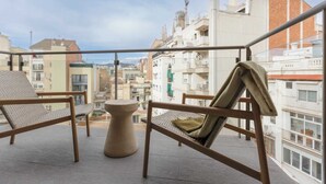 A balcony with two wicker chairs and a small round table, overlooking a cityscape with buildings and a crane.