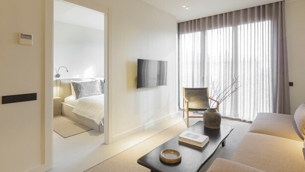 A modern hotel room with a large bed, a seating area with a sofa and a coffee table, a television mounted on the wall, and a large window with curtains.