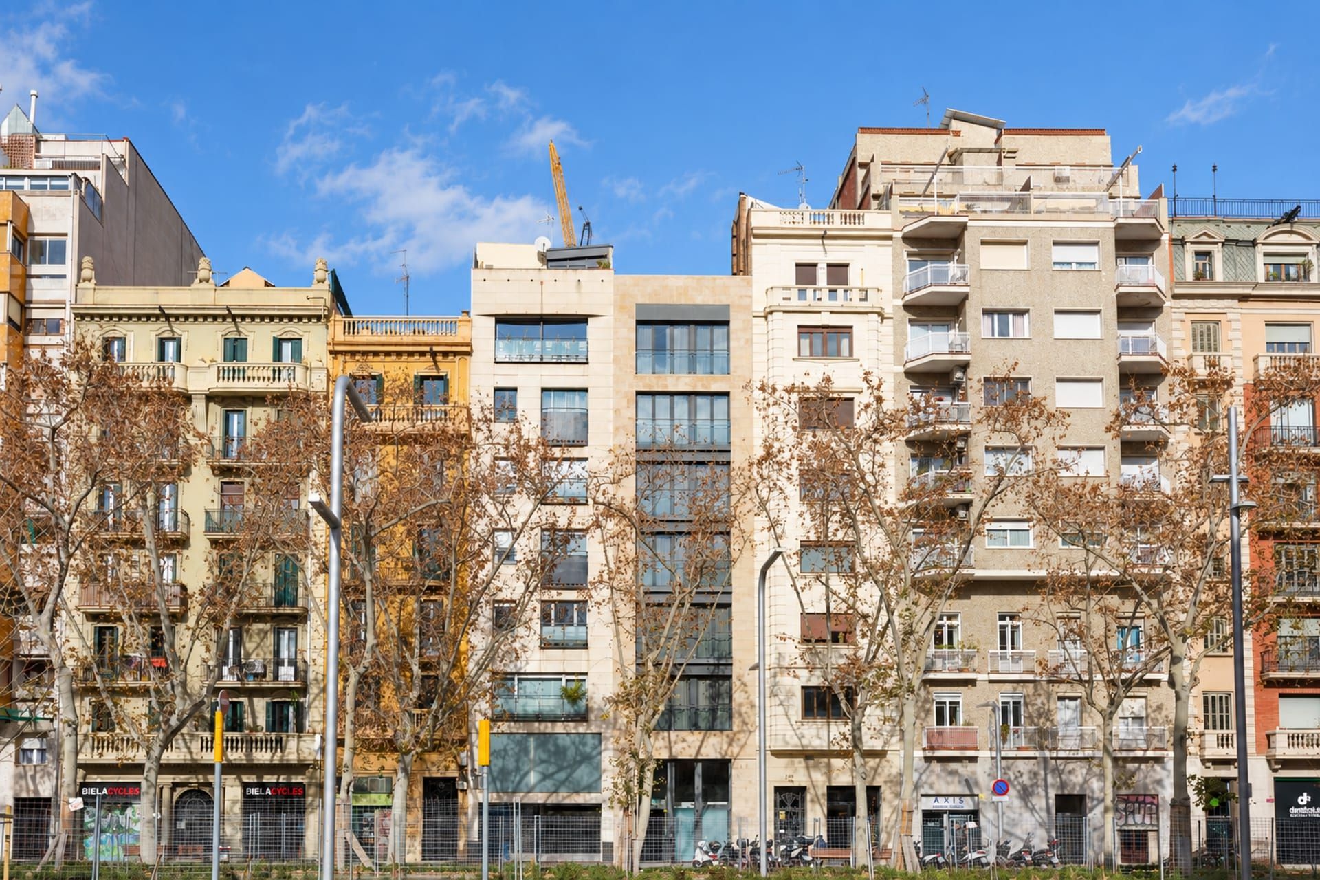 A row of multi-story residential buildings with balconies, a street with trees, and a clear blue sky.