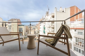 A balcony with two wicker chairs and a small table, overlooking a cityscape with buildings and a clear sky.