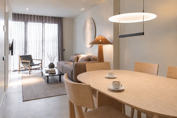 A modern living room with a sofa, a coffee table, and a dining table with chairs. There is a lamp on the table and a large window with curtains.