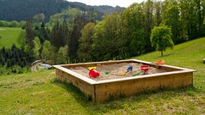 A sandbox with toys and a dirt path in a grassy area with trees and hills in the background. - Apartment 'Schwarzwaldliebe' with Mountain View, Private Terrace and Wi-Fi (Oberharmersbach)