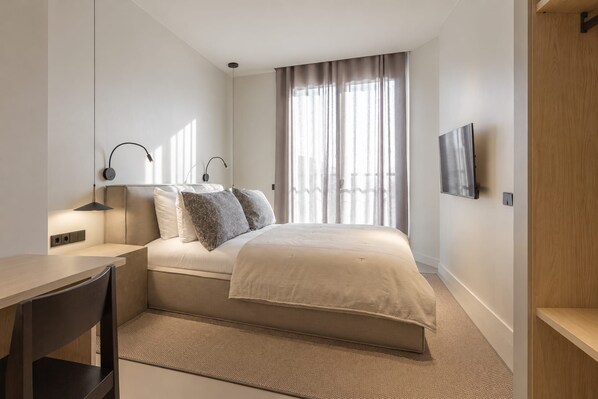 Un dormitorio con cama, mesita de noche, lámpara y televisor instalado en la pared.