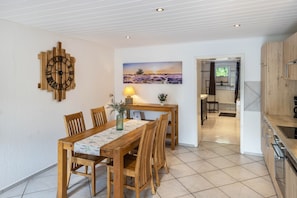 A dining area with a wooden table and chairs, a wall clock, a painting, and a kitchen visible in the background. - Apartment 'An Der Wublitz' with Shared Terrace, Shared Garden and Wi-Fi (Werder (Havel))