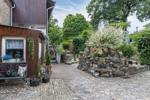 A cobblestone courtyard with a wooden building, stone wall, and various potted plants.