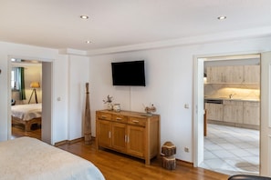 A bedroom with a bed, a wooden dresser, a flat-screen TV, and a kitchen visible through an open door.