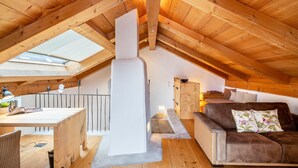 Ein gemütliches Dachzimmer mit Holzbalkendecke, einem Oberlicht, einem Esstisch mit Stühlen, einer Couch und einem Kamin.