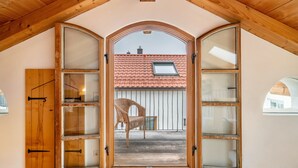 Ein Dachzimmer mit geneigter Decke, Holzbalken, einem großen Bogen und Blick auf ein Gebäude mit roten Ziegeldächern.