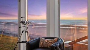 A room with a telescope on a tripod, a leather chair, and a large window overlooking a beach at sunset.