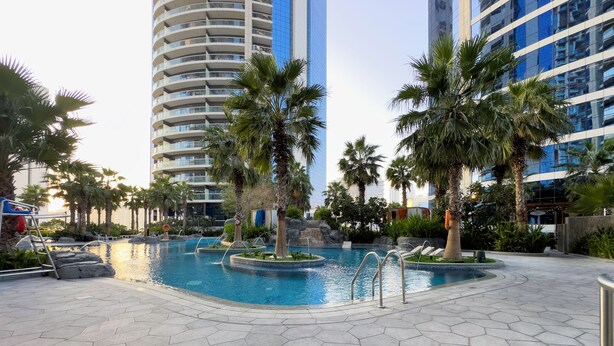 A pool area with hexagonal tiles, palm trees, and modern buildings in the background. - Glamorous 2BR in Damac Paramount Tower (Dubai)