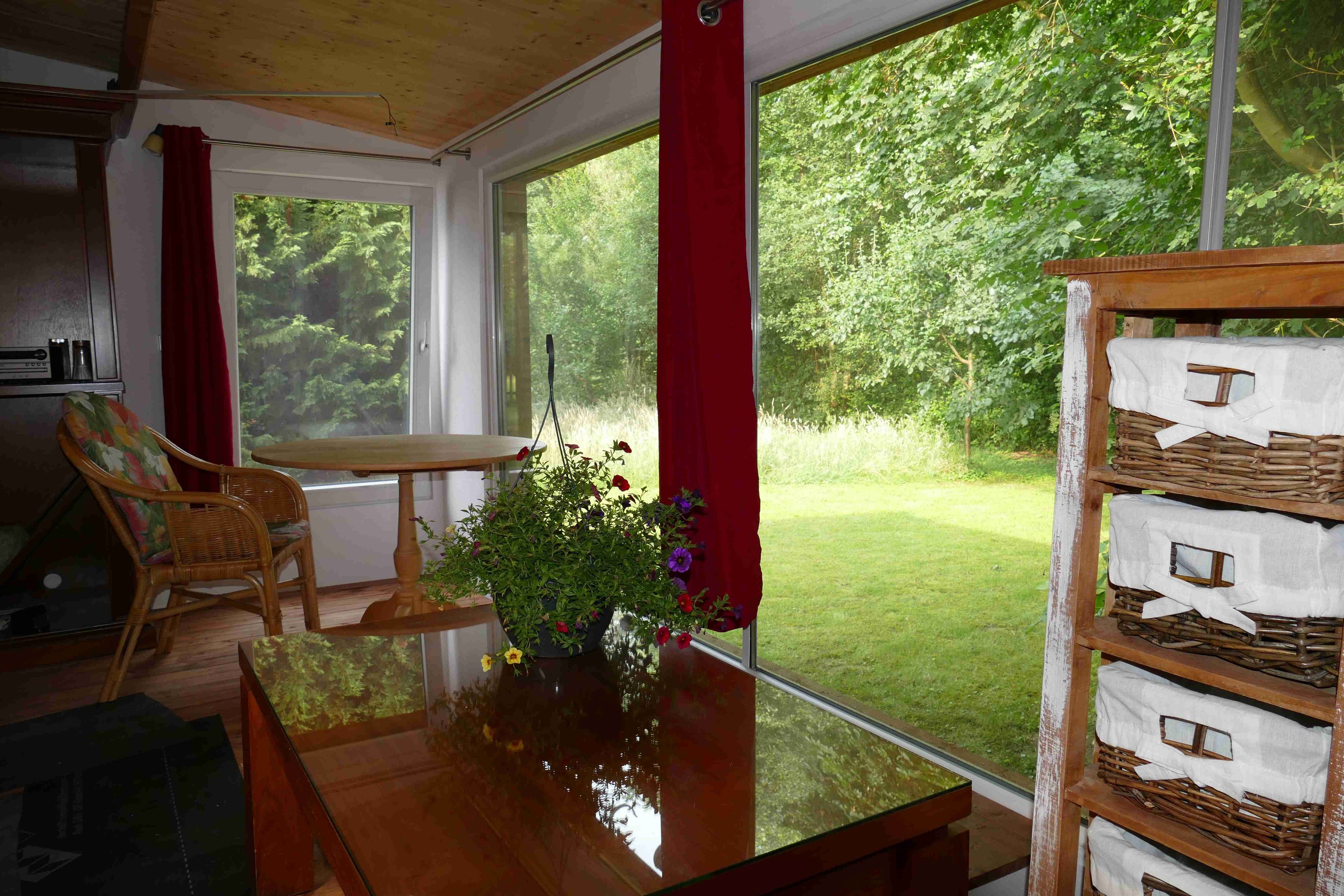Quarto com mesa de madeira, cadeira de vime, planta em vaso com flores e vista para um jardim gramado através de grandes janelas.