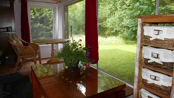 Ein Zimmer mit einem Holztisch, einem Korbstuhl, einem Blumentopf mit Blumen und Blick auf einen grünen Garten durch große Fenster.