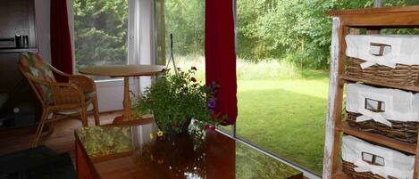 Quarto com mesa de madeira, cadeira de vime, planta em vaso com flores e vista para um jardim gramado através de grandes janelas.