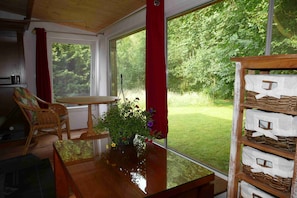 A room with a wooden table, a wicker chair, a potted plant with flowers, and a view of a grassy garden through large windows.