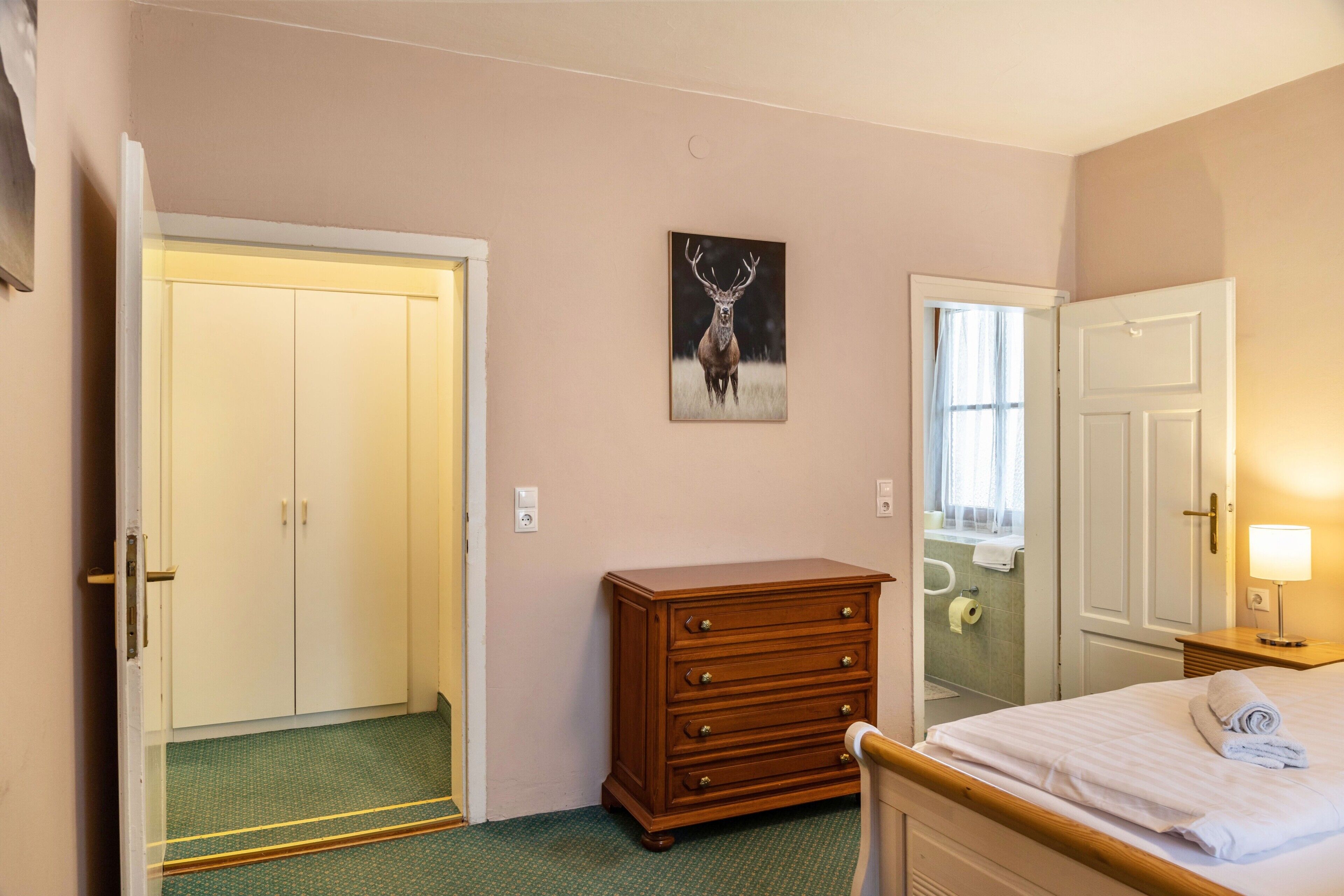 Une chambre d’hôtel avec un lit, une commode en bois, une porte, un miroir et un tableau accroché au mur.