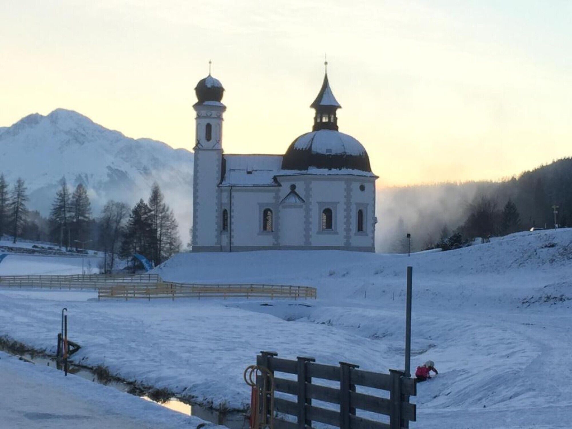 Una iglesia con dos cúpulas y un campanario, rodeada por un paisaje nevado y montañas.