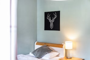 Un lit avec une tête de lit en bois, un oreiller gris, un mur noir orné d’une œuvre représentant une tête de cerf, et une lampe de chevet.