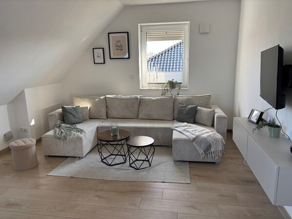 Een moderne woonkamer met een hoekbank, een ronde salontafel en een aan de muur bevestigde televisie.