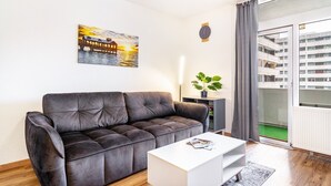A modern living room with a dark brown sofa, a white coffee table, a painting of a sunset, a potted plant, and a view of a building through a glass door.