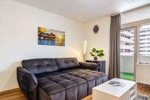 Een woonkamer met een donkerbruine bank, een witte salontafel, een schilderij van een zonsondergang boven een brug, een potplant en uitzicht op een gebouw door een raam.