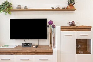 Een moderne woonkamer met een flatscreen-televisie, een houten bureau, witte kasten, decoratieve objecten en een plant.